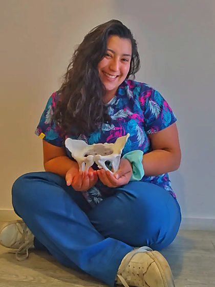 Brenda sentada en el suelo, sonriendo, sosteniendo con las dos manos un modelo anatómico de una pelvis.